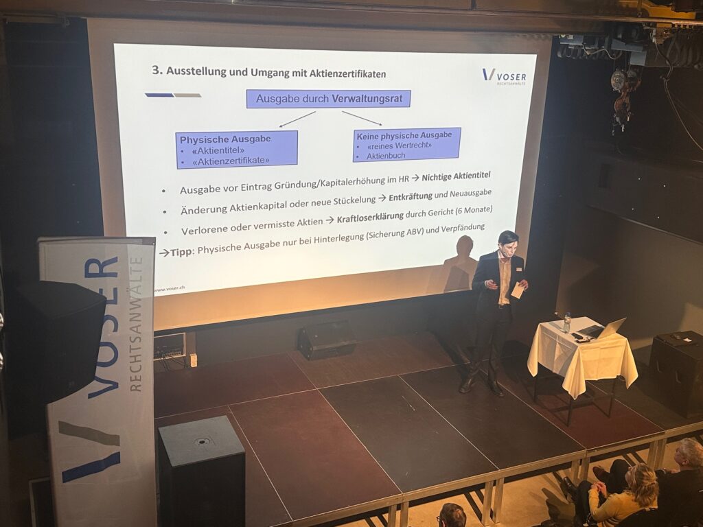 Projected slide from a German presentation about stock certificates, showing a diagram with 'Ausgabe durch Verwaltungsrat' and two branches: 'Physische Ausgabe' and 'Keine physische Ausgabe'. Audience seated in front.