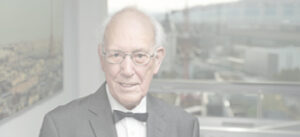 Smiling elderly man in a suit and bow tie seated indoors with a cityscape visible through large windows behind him.