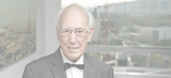 Smiling elderly man in a suit and bow tie seated indoors with a cityscape visible through large windows behind him.