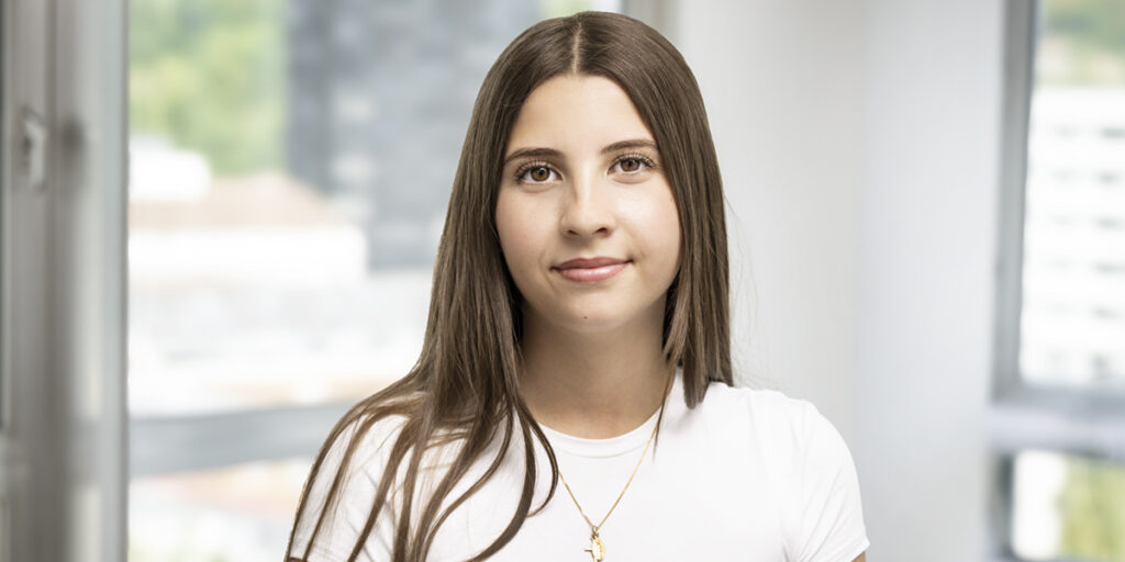 Head-and-shoulders portrait of a young woman with long brown hair in a white shirt, standing by a window.