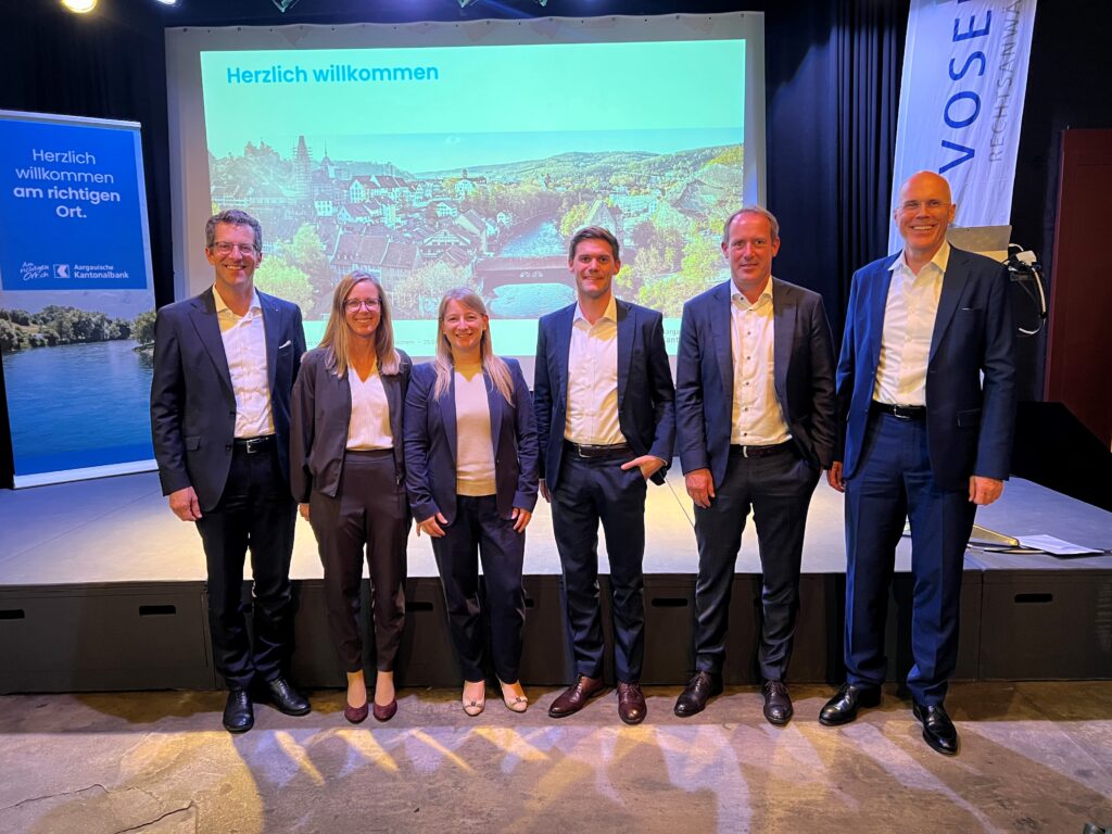 Six professionals in suits stand on a stage for a group photo in front of a projected "Herzlich willkommen" welcome screen and banners in a conference setting, smiling at the camera.
