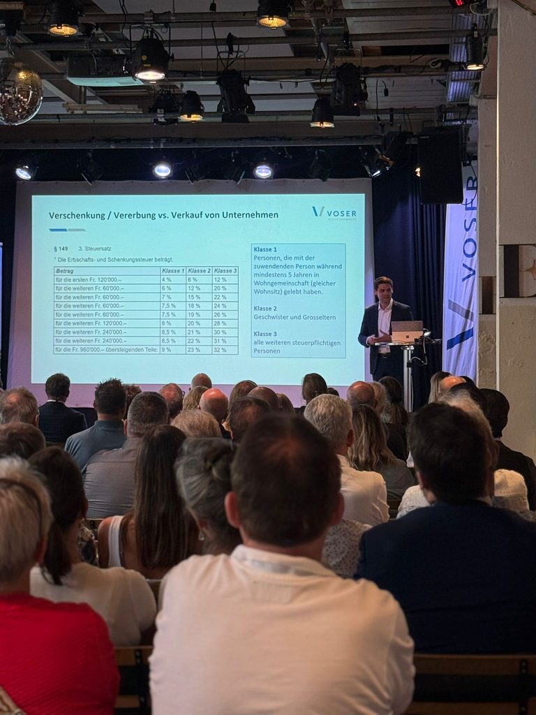 Presenter at a podium giving a slide presentation on stage; projector screen shows German tax/finance content and a Voser logo, audience seated nearby.
