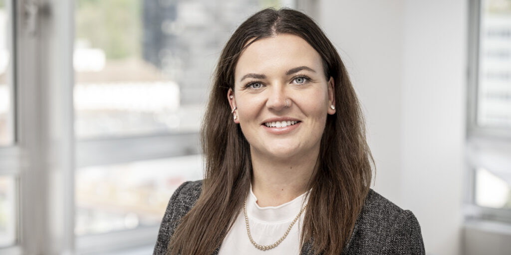 Portrait of a smiling woman with long brown hair, wearing a white blouse and gray blazer in an office setting.