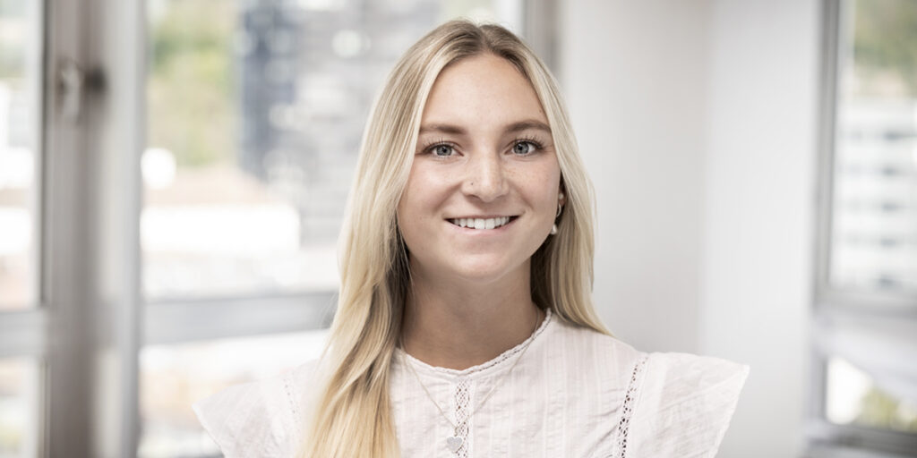 Smiling blonde woman in a white blouse posing indoors with large windows behind her (staff portrait).